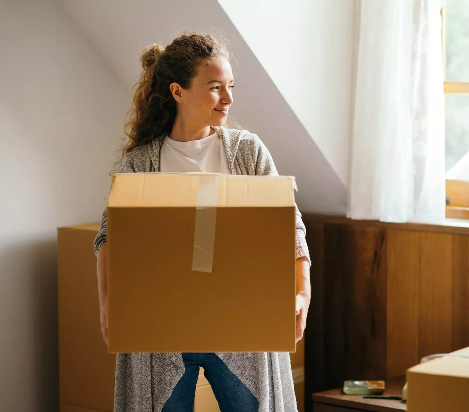 Leende kvinna bär flyttkartong i solbelyst rum under kontorsflytt med Joyweek