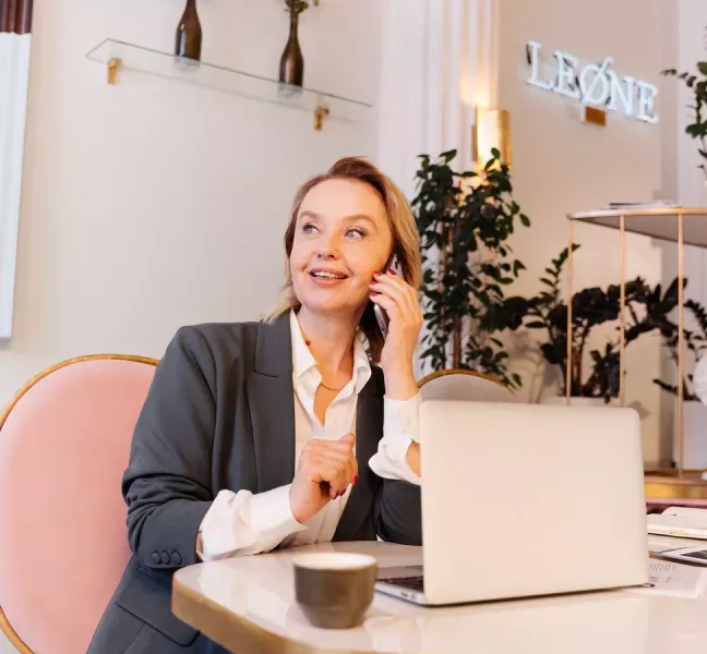 Kvinna i kostym som pratar i telefon vid ett skrivbord med en laptop och en kopp kaffe